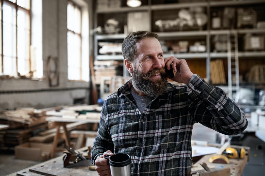 Happy mature male carpenter making phone call during coffee break indoors in carpentery workshop